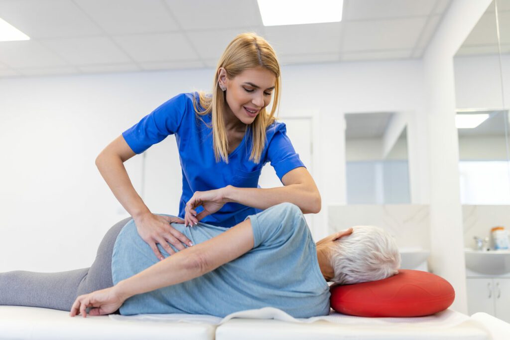Young female chiropractor fixing a lying senior woman's back with hand movements during an occupational health clinic visit.